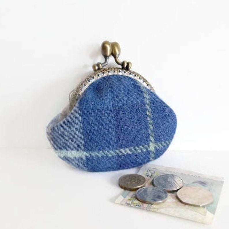 May include: A blue plaid coin purse with a metal clasp, featuring a brass-colored closure. The purse is made of a textured fabric and is accompanied by several coins and a small bill.