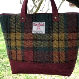May include: A burgundy tote bag with a cork bottom and a plaid pattern in shades of green, red, yellow, and brown. The bag has a label that reads "HAND WOVEN CLOTH Harris Tweed HARRIS TWEED OUTER HEBRIDES OF SCOTLAND 100% PURE NEW WOOL".