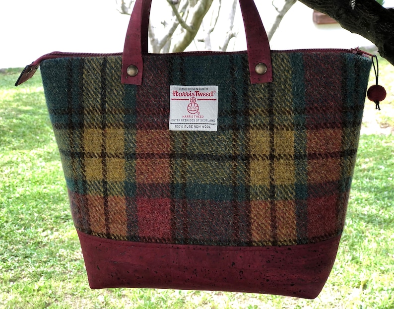 May include: A burgundy tote bag with a cork bottom and a plaid pattern in shades of green, red, yellow, and brown. The bag has a label that reads "HAND WOVEN CLOTH Harris Tweed HARRIS TWEED OUTER HEBRIDES OF SCOTLAND 100% PURE NEW WOOL".