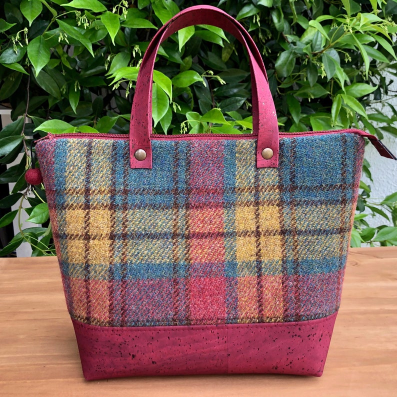 May include: A red, blue, and yellow plaid tote bag with a cork bottom. The bag has two leather handles and a zipper closure.