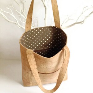 May include: A cork tote bag with a brown cork exterior and a white polka dot fabric lining. The bag has two long straps and a zippered pocket on the front.