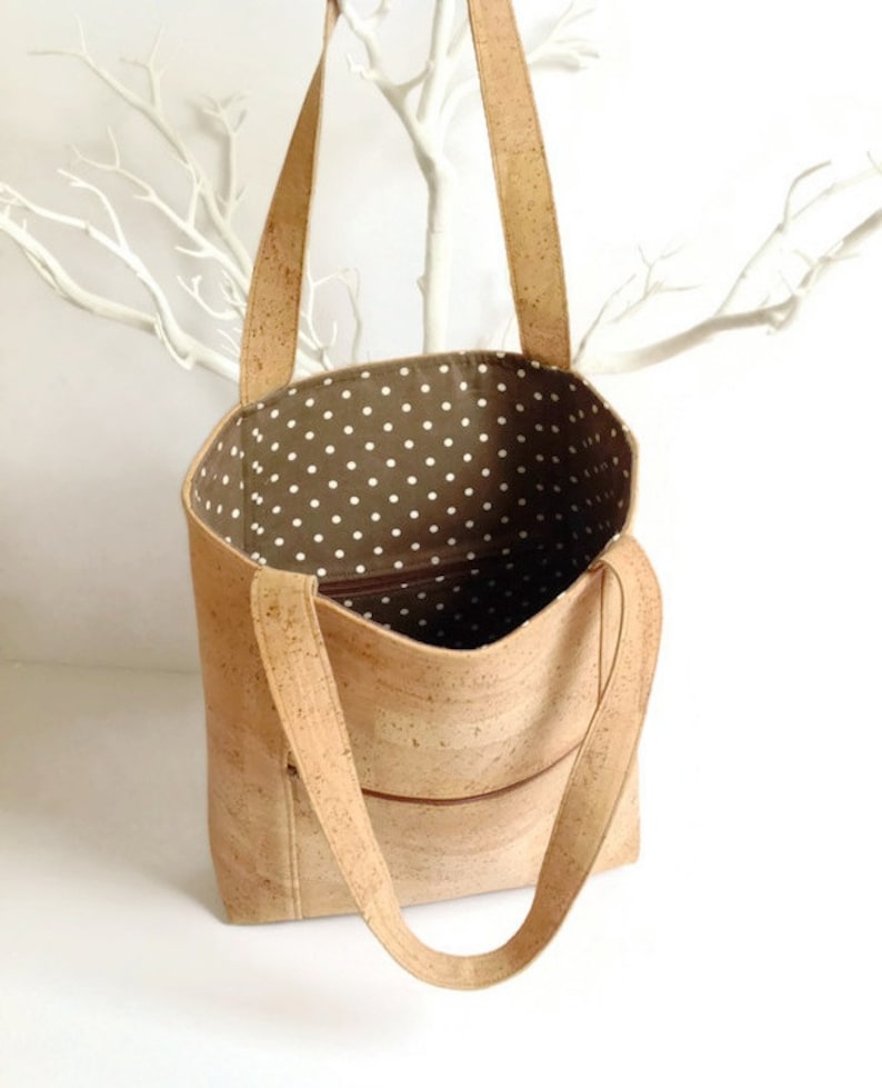 May include: A cork tote bag with a brown cork exterior and a white polka dot fabric lining. The bag has two long straps and a zippered pocket on the front.