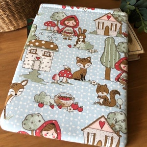 May include: A blue fabric book cover with a whimsical woodland scene. The cover features a red hooded girl, a fox, a squirrel, a rabbit, and two houses with red roofs. The fabric is decorated with a white polka dot pattern.