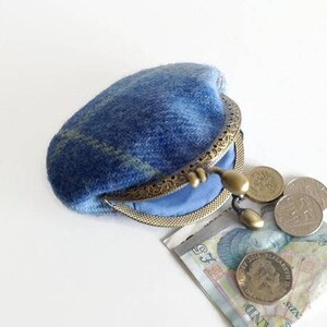 May include: A blue wool coin purse with a brass clasp, open to reveal a light blue interior. Several coins and a paper bill are next to the purse. The purse has a plaid pattern in shades of blue and green.