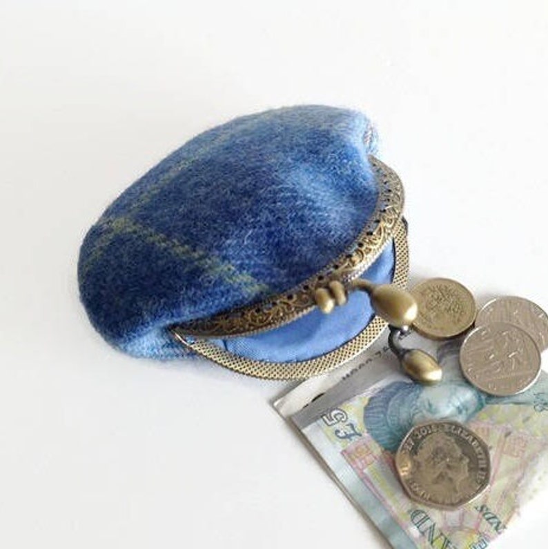 May include: A blue wool coin purse with a brass clasp, open to reveal a light blue interior. Several coins and a paper bill are next to the purse. The purse has a plaid pattern in shades of blue and green.
