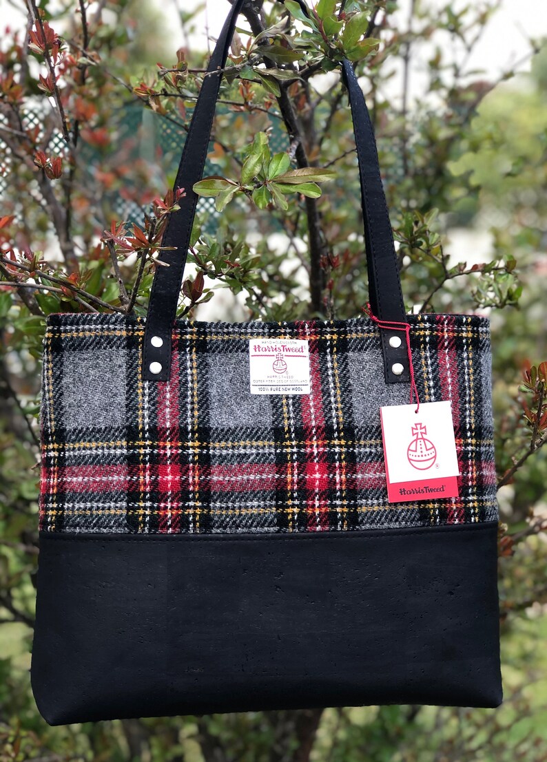 May include: A black tote bag with black straps and a red, yellow, and gray plaid pattern. The bag has a black leather bottom and a label that says "Harris Tweed" with a red and white logo.