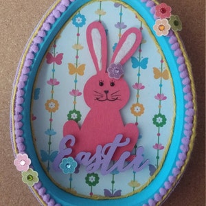 May include: Easter-themed decorative egg-shaped frame. The frame has a light blue border, adorned with purple pom-poms and yellow trim. Inside, a pink bunny with a flower sits above the word "Easter". The background features butterflies and flowers.