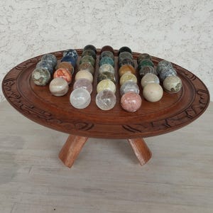 May include: A collection of polished stone spheres in various colors, arranged on a carved wooden tray with three legs. The spheres include clear, pink, beige, and speckled stones. The tray is a rich brown color.