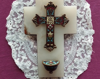 May include: A white marble cross with an ornate, multi-colored metal cross overlay. The metal cross features red, blue, and gold accents. A small, matching basin is below. The cross is set on a white lace doily.