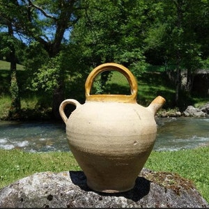 May include: A beige ceramic water jug with a rounded body, a spout, and a handle. The handle and the rim are a darker yellow color. The jug is sitting on a rock in front of a river and green foliage.