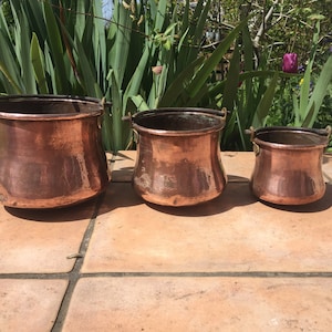 Puede incluir: Tres ollas de cobre antiguas de diferentes tamaños. Cada olla tiene un cuerpo redondeado, un borde oscuro y pequeñas asas de metal. El cobre tiene un tono marrón rojizo cálido, con pátina visible. Las ollas están dispuestas sobre una superficie de baldosas de terracota.