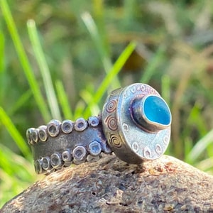 May include: A silver ring featuring a blue glass stone set in a circular bezel. The band is adorned with a row of small circles.