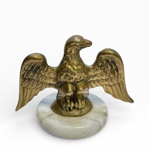 May include: A brass eagle sculpture with outstretched wings, mounted on a round, marble-like base. The eagle is detailed with textured feathers and a prominent beak. The sculpture is a decorative item.