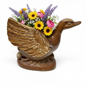 May include: A brown ceramic duck-shaped planter featuring a glossy finish and molded wing details. The planter is filled with an arrangement of artificial flowers, including yellow sunflowers, purple lavender sprigs, and small pink blossoms.