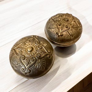 May include: Two antique bronze door knobs with ornate floral and scrollwork designs. The knobs are round with a slightly aged appearance, suggesting they are vintage or vintage-inspired. The detailed carvings add a touch of elegance.