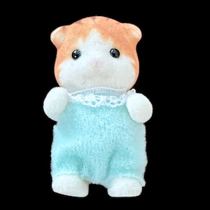 Op de afbeelding: Een klein, pluche hamsterspeelgoed met oranje en witte vacht. De hamster draagt een lichtblauwe onesie met witte rand en witte poten. Het heeft zwarte ogen en een roze neus.