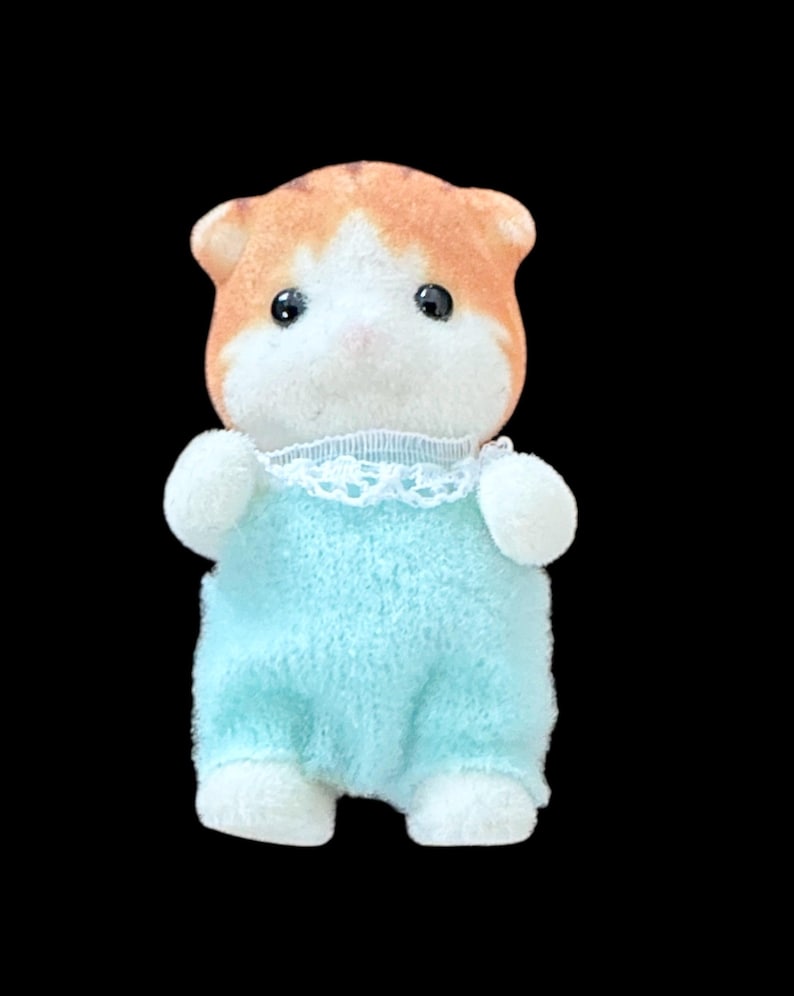 Op de afbeelding: Een klein, pluche hamsterspeelgoed met oranje en witte vacht. De hamster draagt een lichtblauwe onesie met witte rand en witte poten. Het heeft zwarte ogen en een roze neus.