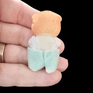 Op de afbeelding: Een kleine, vintage poppenfiguur, van achteren bekeken. De pop heeft een perzikkleurig hoofd en draagt een witte top met kanten rand en lichtblauwe shorts. De pop wordt in een hand gehouden tegen een zwarte achtergrond.