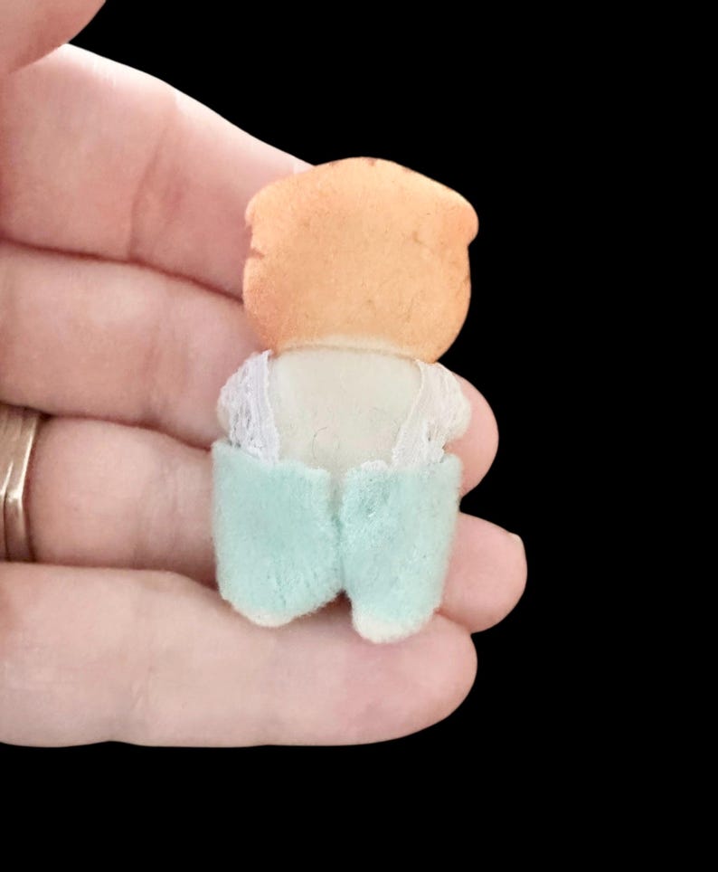 Op de afbeelding: Een kleine, vintage poppenfiguur, van achteren bekeken. De pop heeft een perzikkleurig hoofd en draagt een witte top met kanten rand en lichtblauwe shorts. De pop wordt in een hand gehouden tegen een zwarte achtergrond.