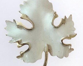 May include: A vintage leaf-shaped brooch with a white enamel finish and gold-tone metal accents. The brooch features a detailed design with a stem and curved accents, creating a classic and elegant look. The leaf design is a timeless accessory.