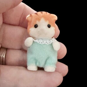 Op de afbeelding: Een miniatuur oranje en witte kattenfiguur met een lichtblauwe jumpsuit met een witte kanten kraag. De kat heeft zwarte ogen en witte poten. Het beeldje wordt in een hand gehouden tegen een zwarte achtergrond.