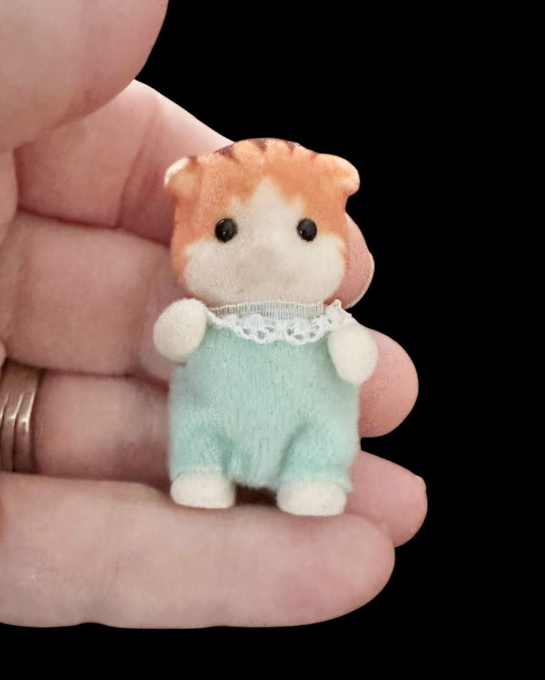 Op de afbeelding: Een miniatuur oranje en witte kattenfiguur met een lichtblauwe jumpsuit met een witte kanten kraag. De kat heeft zwarte ogen en witte poten. Het beeldje wordt in een hand gehouden tegen een zwarte achtergrond.