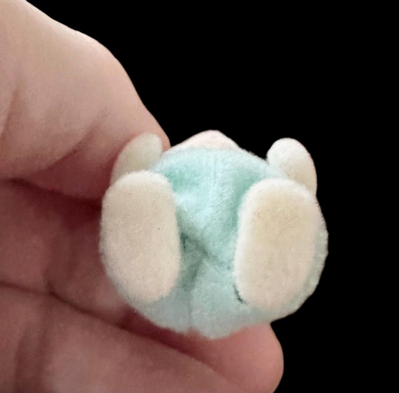 Op de afbeelding: Een klein, pluche speelgoed in de vorm van een baby, van onderaf gezien. Het speelgoed heeft een lichtblauwe body en gebroken witte voeten en handen. De achtergrond is effen zwart.