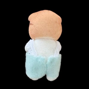 Op de afbeelding: Een vintage pop met een perzikkleurig hoofd en een witte top met kanten rand. De pop draagt lichtblauwe shorts. De pop staat tegen een zwarte achtergrond.