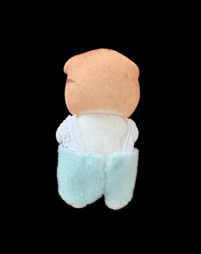 Op de afbeelding: Een vintage pop met een perzikkleurig hoofd en een witte top met kanten rand. De pop draagt lichtblauwe shorts. De pop staat tegen een zwarte achtergrond.