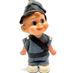 May include: A small doll wearing a grey felt uniform with a black sash, a matching grey hat, and white shoes. The doll has blue eyes, rosy cheeks, and silver star-shaped accents on the shoulders of the uniform. A red detail is visible on the chest.