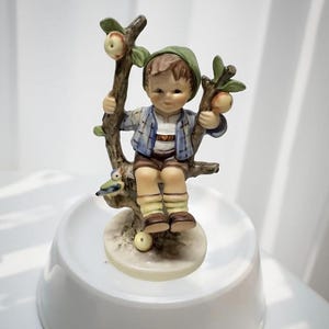 May include: A porcelain figurine of a young boy seated on a tree branch, holding onto the branches. The boy wears a green hat, blue jacket, brown shorts, and yellow socks. The tree has apples and a small bird. The figurine is on a white base.