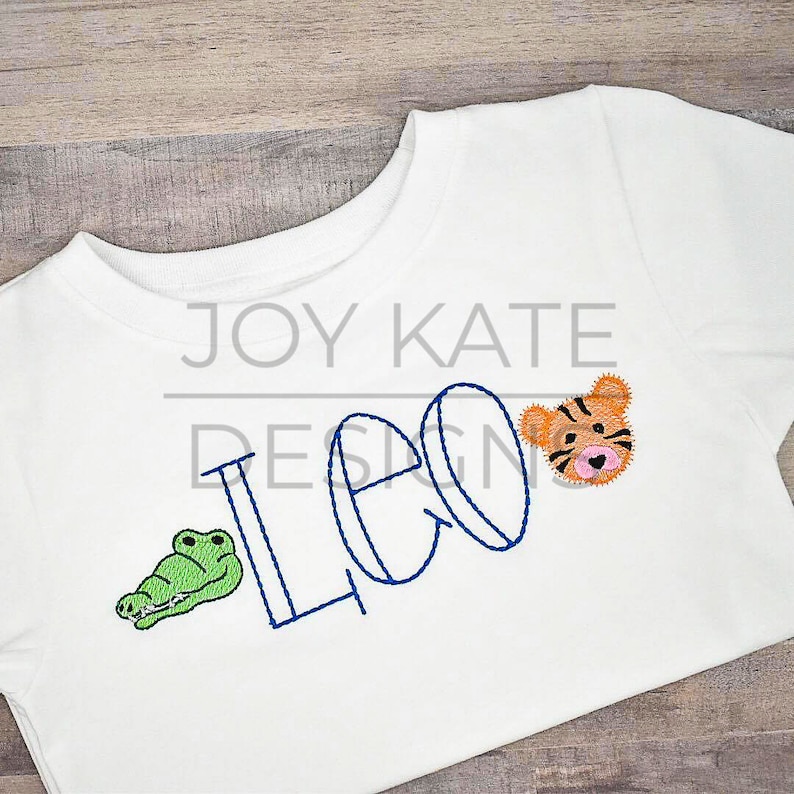 以下が含まれることがあります： 白いTシャツに青い糸で「レオ」という名前が刺繍されています。名前の両側に緑色のワニとオレンジ色の虎の顔が刺繍されています。