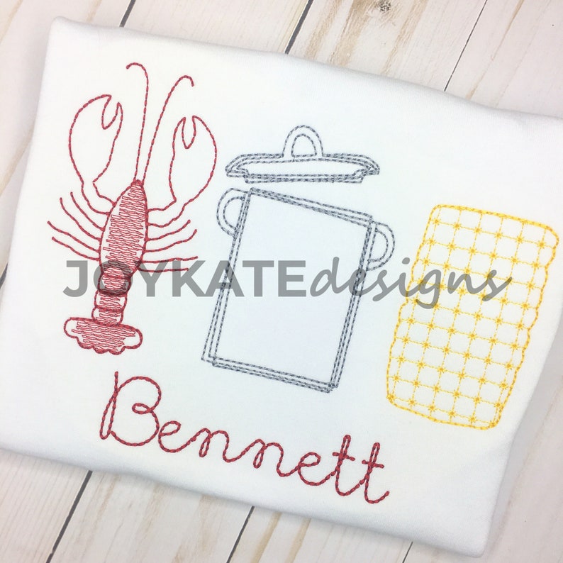 May include: Embroidered design of a red lobster, a gray pot with a lid, and a yellow corn cob with the name "Bennett" in red cursive.