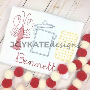 May include: Embroidered white t-shirt with a red lobster, a gray pot with a lid, and a yellow checkered pattern. The name "Bennett" is embroidered in red below the design.
