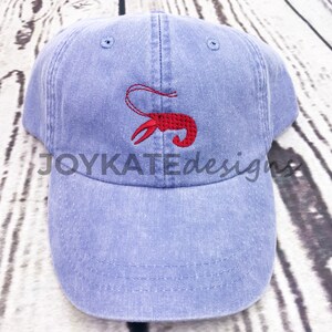 May include: A blue denim baseball cap with a red embroidered lobster design. The cap has a curved brim and an adjustable strap.