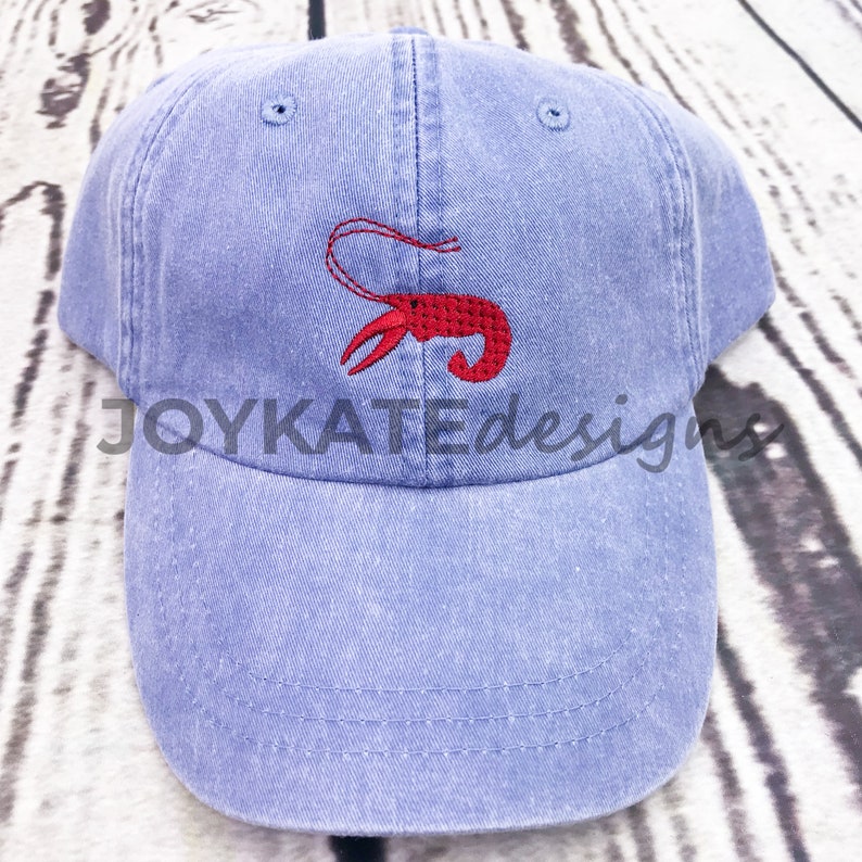 May include: A blue denim baseball cap with a red embroidered lobster design. The cap has a curved brim and an adjustable strap.