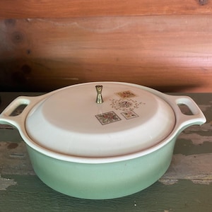 May include: A round, light green casserole dish with a matching white lid. The lid features a central metallic knob and three square decorative patterns depicting floral and botanical motifs in muted earth tones. The dish has two integrated handles.