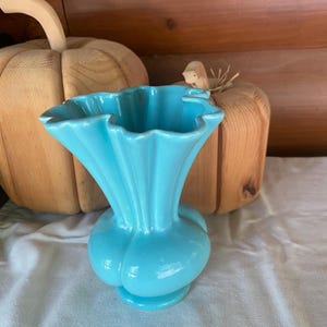 May include: A bright turquoise ceramic vase with a bulbous base and a wide, ruffled, fluted rim. The vase features a smooth, glossy finish and a vertical ribbed design that extends from the neck to the base, creating a distinct scalloped edge.
