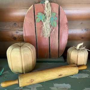May include: Wooden fall-themed decorations including a large pumpkin cutout, two smaller pumpkin-shaped objects, and a wooden rolling pin. The large pumpkin is painted orange with a green stem and raffia. The rolling pin is light brown.