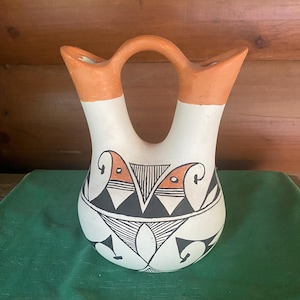 May include: A ceramic wedding vase with two spouts connected by a central handle. The vessel features a cream-coloured base with hand-painted geometric patterns in black and terracotta orange, including triangular and linear motifs around the body.