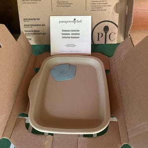 Op de afbeelding: Een rechthoekige beige stenen bakvorm met een lichtblauwe plastic schraper erin, verpakt in een kartonnen doos met een Pampered Chef instructieboekje. De doos bevat tekst over de veiligheid voor gebruik in de oven en vaatwasser.