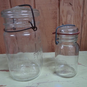 Puede incluir: Dos tarros de vidrio transparentes con tapas de metal. El tarro más grande tiene un cierre de alambre con pestillo. El tarro más pequeño tiene una junta de goma roja.