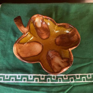 Könnte beinhalten: Eine Keramikschale in Form eines vierblättrigen Kleeblatts mit glänzender Oberfläche. Die Schale zeigt einen Farbverlauf aus warmen Braun- und Goldgelbtönen, mit dunkelbraunen Akzenten in der Mitte jedes Blattsegments und einem kleinen Stiel.