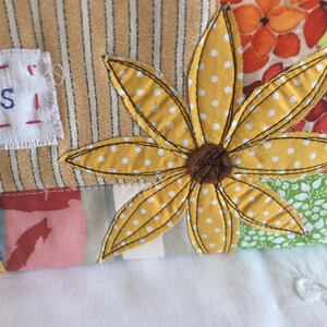 May include: A yellow fabric flower with white polka dots is sewn onto a patchwork fabric background. The fabric background includes stripes, floral patterns, and a white label with the word "needles" stitched in red thread.