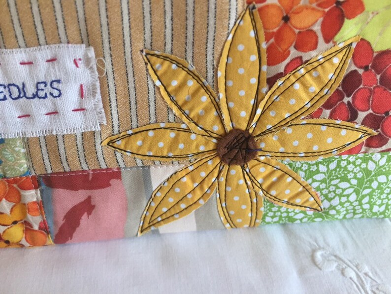 May include: A yellow fabric flower with white polka dots is sewn onto a patchwork fabric background. The fabric background includes stripes, floral patterns, and a white label with the word "needles" stitched in red thread.