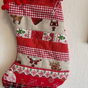 May include: A Christmas stocking constructed from horizontal fabric strips featuring red, white, and beige patterns including reindeer, Santa Claus, gingham, and plaid. The top edge is decorated with red pom-pom trim and a small red ribbon bow.