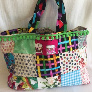 May include: A colourful patchwork tote bag with green pom pom trim. The bag features a variety of fabrics, including floral prints, polka dots, and geometric patterns. The bag has two black fabric handles.