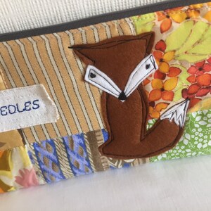 May include: A brown felt fox applique with white felt accents is sewn onto a patchwork fabric pouch. The pouch is made of various patterned fabrics in shades of brown, beige, green, blue, and orange. The pouch has a white label that says "My Needles".