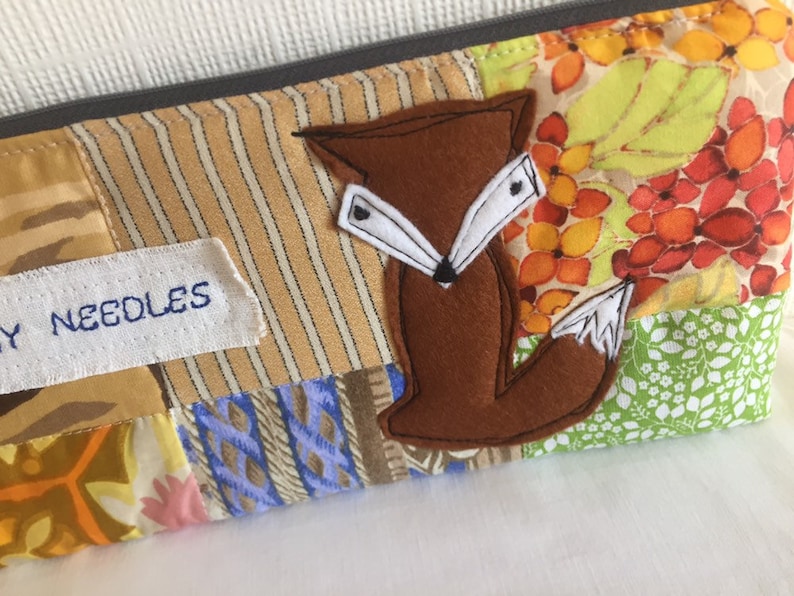 May include: A brown felt fox applique with white felt accents is sewn onto a patchwork fabric pouch. The pouch is made of various patterned fabrics in shades of brown, beige, green, blue, and orange. The pouch has a white label that says "My Needles".