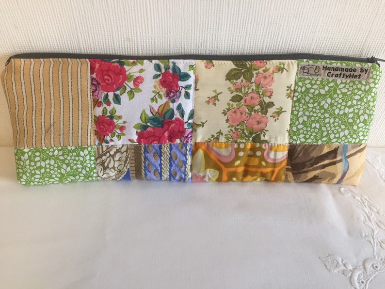 May include: A patchwork fabric zippered pouch with a variety of floral and geometric patterns in shades of green, yellow, pink, blue, and brown. The pouch has a label that reads "Handmade by CraftyHet".